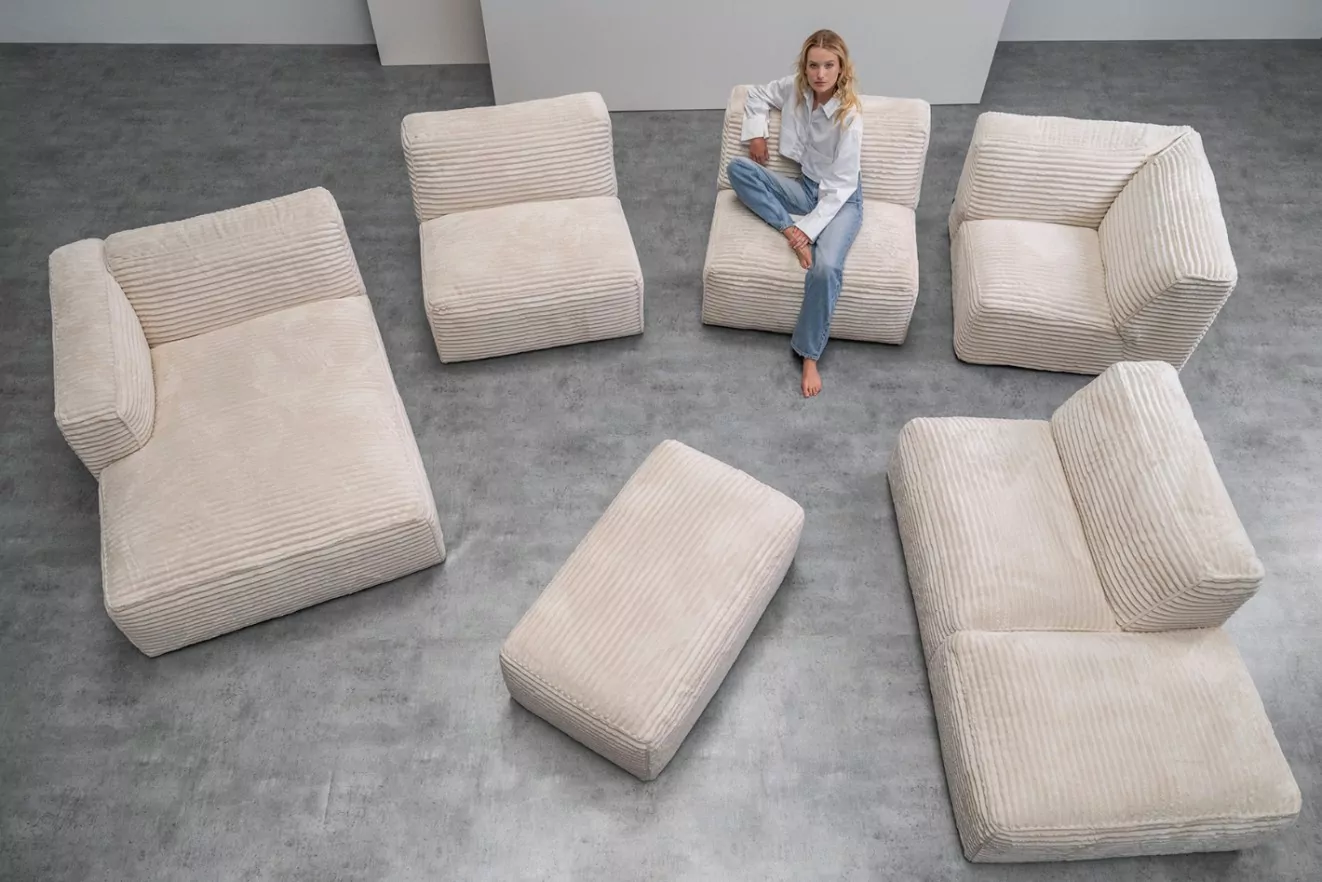 Beige modular corduroy sofa set with multiple seating sections and an ottoman arranged on a grey floor, with a woman sitting on one of the modules. Pastel Perfection for Contemporary Living Spaces