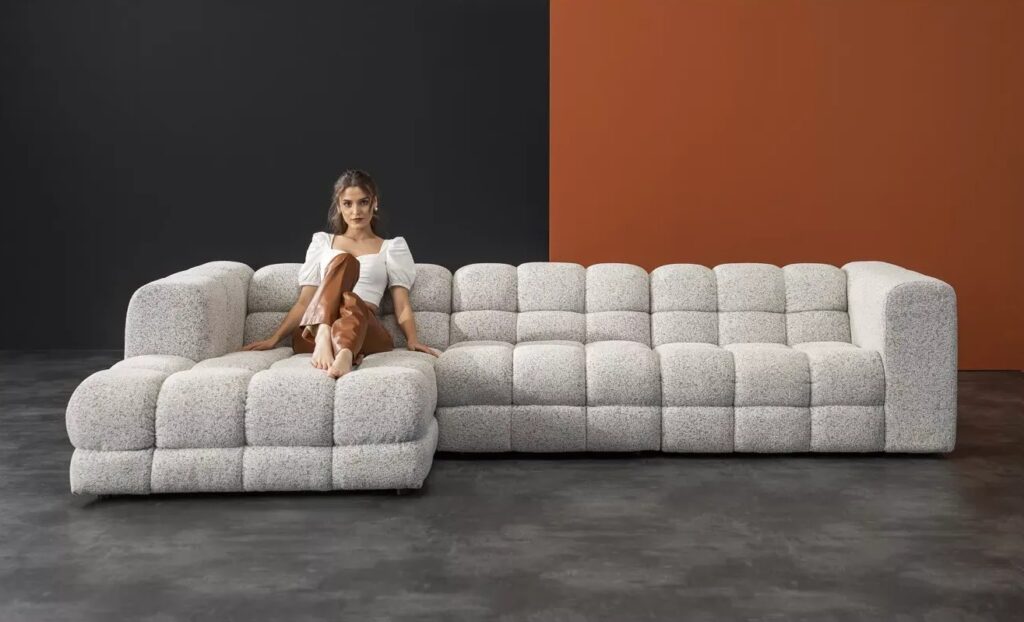 Modern modular corner sofa in light grey boucle fabric with tufted square design, styled in a minimal interior with dark grey floor and two-tone wall in black and terracotta, with a woman sitting casually on the chaise section. Story Sofa