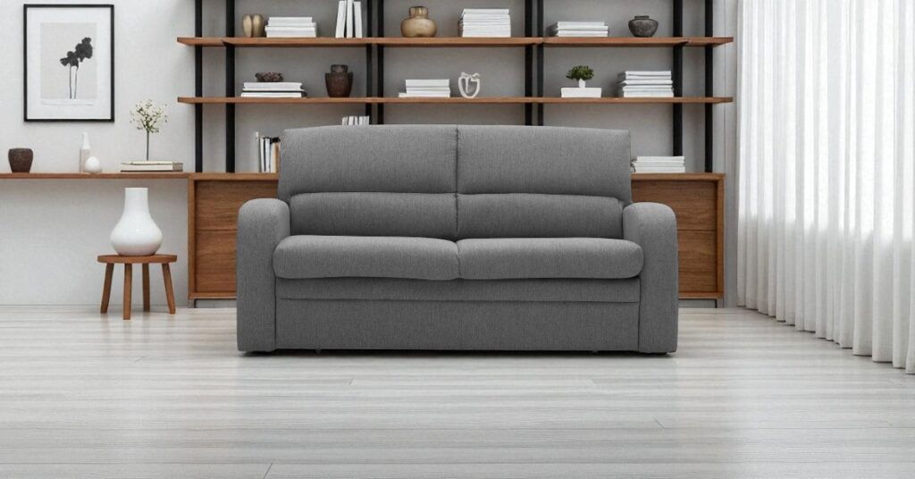 Compact grey two-seater sofa placed in a modern living room with open wooden shelving unit behind, decorated with books, ceramics, and minimalist ornaments, with floor-to-ceiling curtains allowing natural light in.