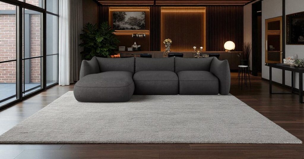 Contemporary living room with a black corner sofa, large grey rug, dark wooden flooring, floor-to-ceiling windows and warm ambient lighting creating a dramatic and elegant interior.