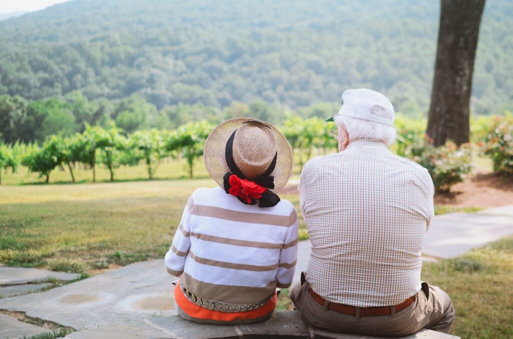 Elderly couple sitting on a stone path, looking out over green vineyards and a scenic hillside on a summer’s day.