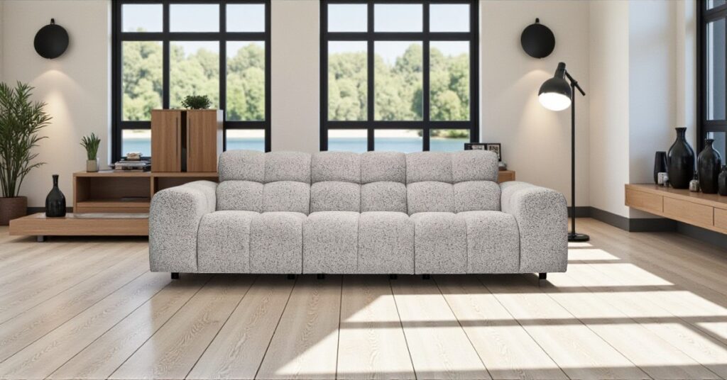 Contemporary living room with a grey textured sofa placed on light wooden flooring, large black-framed windows in the background, modern wooden furniture, plants, and a floor lamp creating a stylish interior.