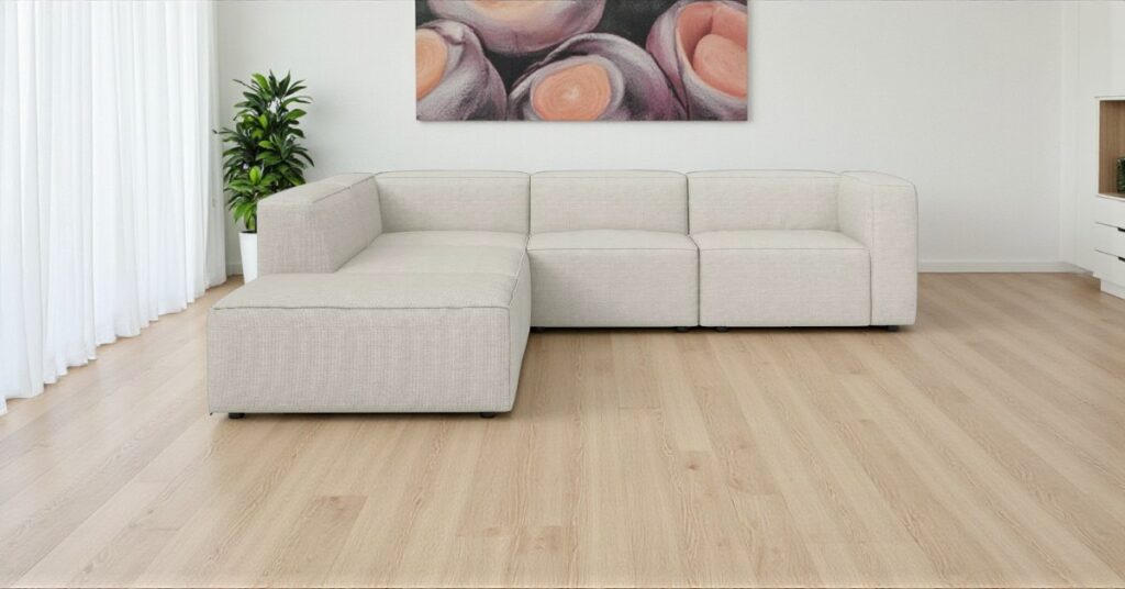Beige modular corner sofa placed against a blank white wall in a modern living room with light wooden flooring, white curtains, a green potted plant, and abstract wall art above the sofa.