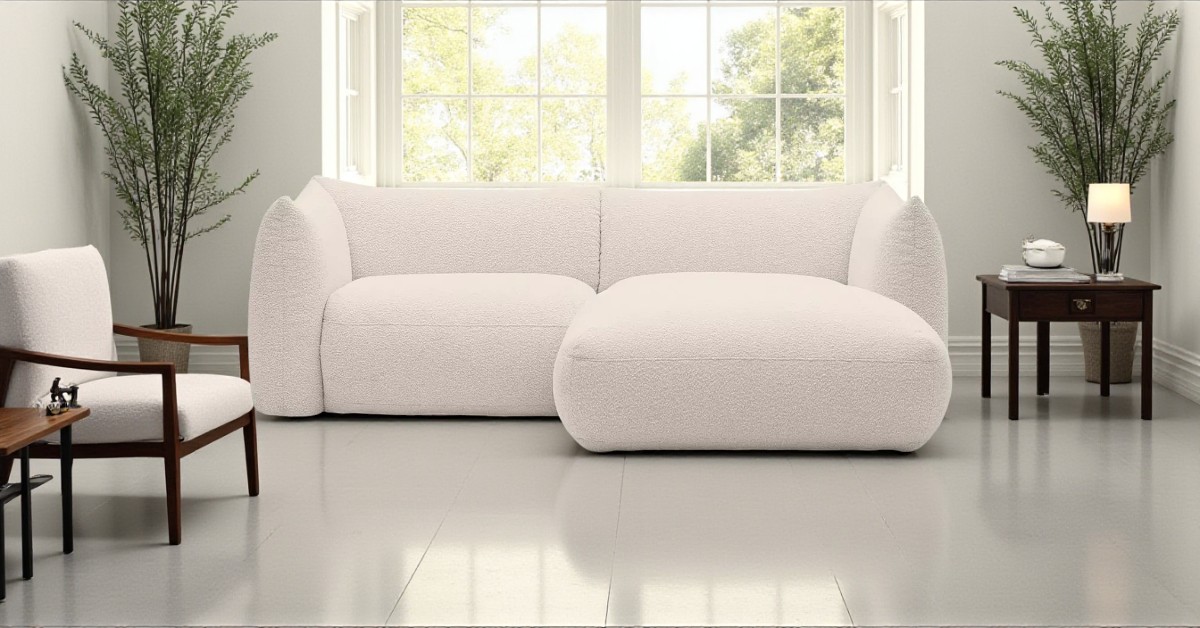 Modern living room with a large bay window, featuring a white boucle corner sofa, wooden armchair, side tables with lamps, and tall potted plants creating a cosy seating area.